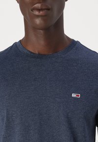 Prim-plan cu o persoană care poartă un tricou bleumarin cu decolteu rotund și un mic logo dreptunghiular roșu, alb și bleumarin pe partea stângă a pieptului.