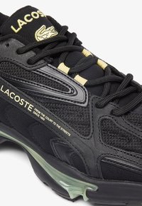 Sneaker nera Lacoste realizzata in materiali mesh e sintetici, con accenti testurizzati, branding dorato e suola trasparente.