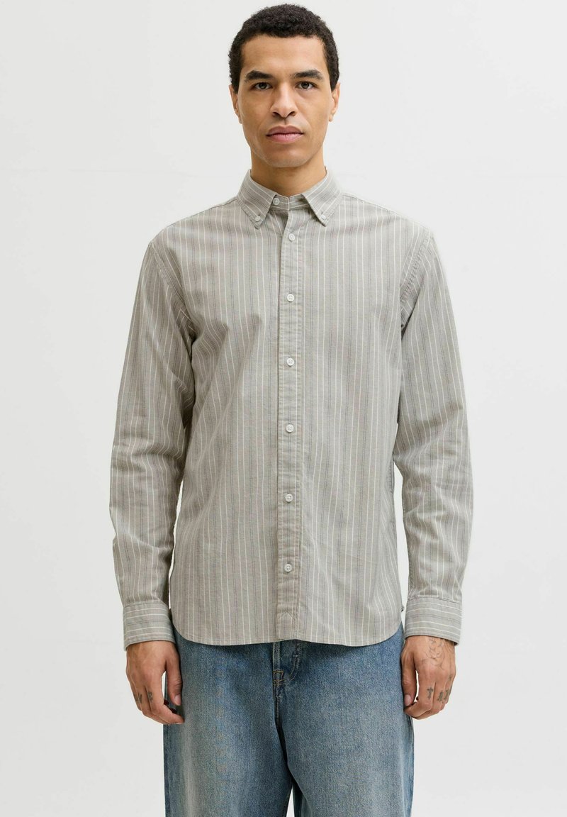 Mladý muž oblečený v zapnuté svetlošedej, vertikálne pruhovanej košeli s dlhým rukávom a voľných modrých džínsach, stojaci proti jednoliatemu bielemu pozadiu.