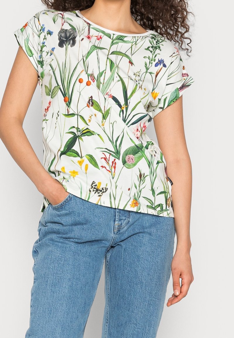 Femme portant une chemise blanche à manches courtes avec un imprimé floral et papillons coloré et un jean bleu en denim, main dans la poche.