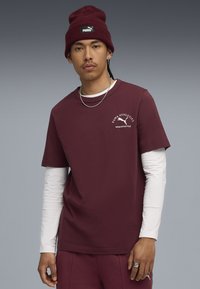 Bordeauxfarbenes T-Shirt, das über einem weißen Langarmshirt getragen wird, mit einem Puma-Logo und Text auf der Brust, ergänzt durch eine passende bordeauxfarbene Mütze.