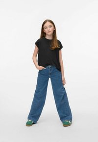 T-shirt noir à manches courtes associé à un jean en denim bleu à jambe large. Le modèle porte des baskets vertes. Tenue simple et décontractée avec une coupe ample.