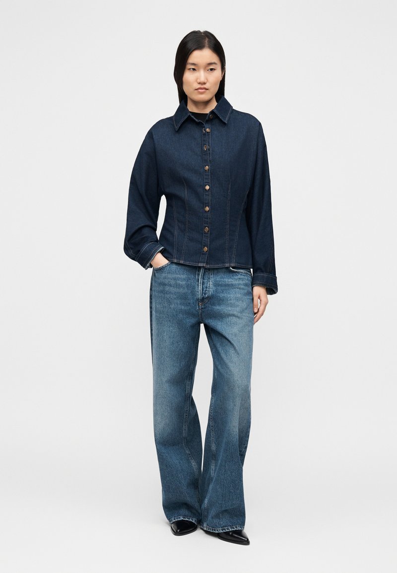 Veste en jean foncé avec boutonnière et col à revers, associée à un jean en denim clair à coupe ample. Texture lisse, sans motifs ni logos visibles.