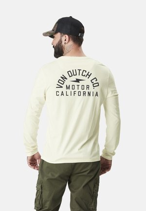 T-shirt à manches longues crème avec un imprimé graphique noir au dos, associé à un pantalon cargo vert olive et une casquette camouflage noire.