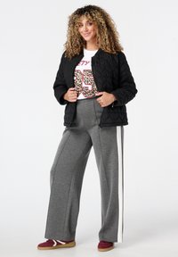 Vrouw met krullend haar in een zwart gewatteerd jasje, een witte grafische t-shirt, grijze wijde broek met een witte streep en kastanjebruine sneakers.