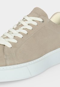 Μπεζ καστόρινο χαμηλό sneaker με λευκά κορδόνια και χοντρή λευκή ελαστική σόλα, με μεταλλικά κορδόνια και ορατές λεπτομέρειες ραφής.
