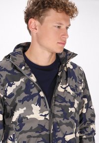 Giubbotto mimetico con motivi grigi, blu navy e beige. Presenta un colletto alto e una zip frontale. Indossato sopra una maglietta blu navy a girocollo.