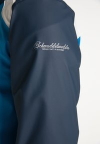 Marinblå vattentät jacka med texturerad yta, kontrasterande blå detaljer och silverbroderad logotyp som står 'Schmuddelwedda' på ärmen.