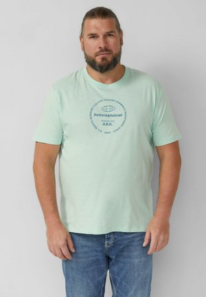 Uomo con la barba e capelli lunghi raccolti indossa una maglietta verde chiaro con la scritta circolare "Rethink&Reload" e jeans blu, in piedi davanti a una parete bianca.