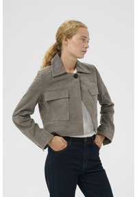 Trumpas pilkas zomšos švarkas su apykakle, vieno mygtuko užsegimu ir dviem priekinėmis kišenėmis, derinamas su tamsių džinsų kelnėmis.