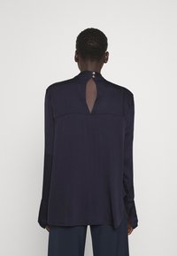 Blusa navy a maniche lunghe con finitura testurizzata, caratterizzata da un'apertura a chiave sul retro e due bottoni sul collo. Taglio dritto e vestibilità ampia.