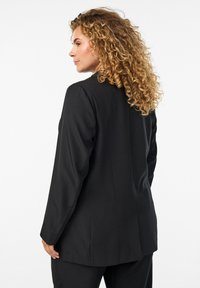 Black tailored blazer featuring smooth fabric, long sleeves, a structured fit, and a single vent at the back, styled with minimal detailing.