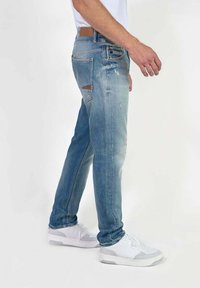 Jean en denim bleu clair avec une coupe décontractée, présentant des détails usés. Associés à des baskets blanches, créant un look décontracté et moderne.