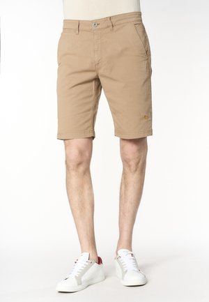 Gambe maschili con pantaloncini beige al ginocchio e scarpe da ginnastica bianche con dettagli rossi, in piedi su uno sfondo bianco semplice.