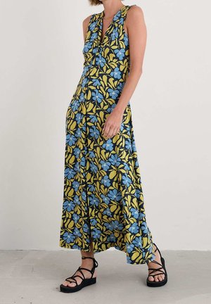 Kvinde iført en ærmeløs lang kjole med blåt og gult blomsterprint og sorte sandaler med stropper mod en ensfarvet lys baggrund.