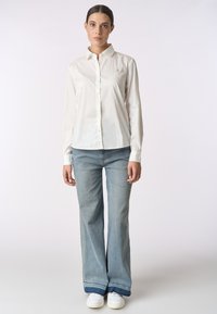 Camicia bianca con bottoni e maniche lunghe, con un piccolo logo ricamato sul petto. Abbinata a jeans svasati blu chiaro e sneakers bianche.