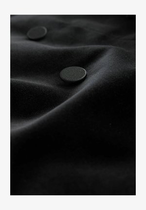 Juodas audinys su minkšta tekstūra, puošiamas dviem iškilusiomis apvaliomis mygtukais su tekstūruota paviršiumi, sukuriantys subtilesnį vizualinį kontrastą.