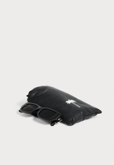 Lunettes de soleil rectangulaires noires reposant sur une pochette noire douce avec un logo de palmier blanc sur un fond clair.