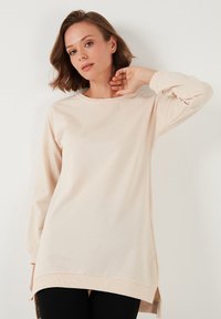Túnica beige de manga larga con escote redondo y aberturas laterales, confeccionada en suave tela de algodón. Presenta detalles acanalados en el bajo y los puños.