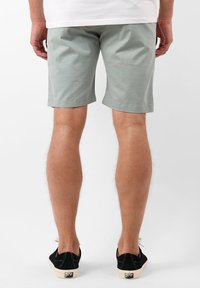 Shorts vert clair en coton, avec un design simple, une coupe droite et des bords ourlés. Portés avec des chaussures noires sur un fond neutre.