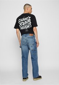 Camiseta de algodón negra con el texto en blanco "CAMBIA TUS PENSAMIENTOS" en la parte de atrás, combinada con pantalones de mezclilla azules con bolsillos tipo cargo.