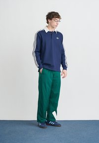 Μπλε ναυτικό πόλο με λευκό γιακά, με τρεις λευκές ρίγες στα μανίκια. Πράσινο φαρδύ παντελόνι, μπλε αθλητικά παπούτσια με τρεις ρίγες.