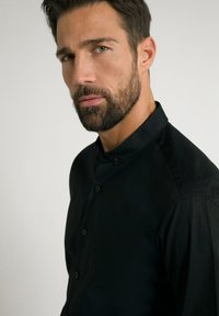 Schwarzes Hemd mit Knöpfen und Kragen, glatte Textur, taillierter Schnitt und keine sichtbaren Muster oder Verzierungen, getragen von einem Mann im Profil.