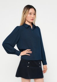 Cămașă albastră închis cu mânecă lungă, cu închidere cu nasturi și umeri bombati, asortată cu o fustă închisă la culoare, cu nasturi argintii și o textură de catifea.