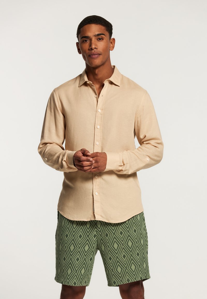Camicia beige a maniche lunghe con chiusura a bottoni e vestibilità rilassata, abbinata a pantaloni corti verdi con un motivo a rombi.