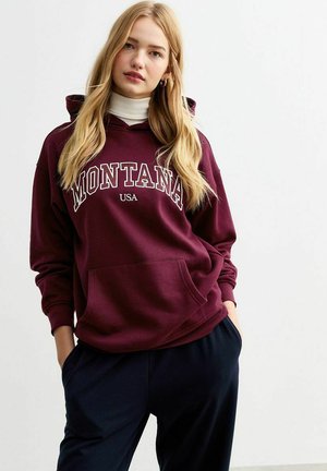 Burgundy hoodie featuring a front kangaroo pocket and white "MONTANA USA" text. Worn over a white turtleneck, paired with navy pants.