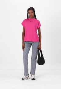 Pink cotton T-shirt with short sleeves and a crew neckline, paired with light gray jeans. Black woven bag and athletic shoes, standing on a plain background.