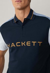 Marineblaues Polohemd mit Blau- und Weißakzenten, das ein goldenes "HACKETT"-Logo trägt. Das Material wirkt glatt, mit einem klassischen Kragen und Knopfdetails.