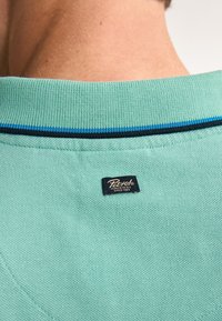 Tyrkysová polo košeľa s textúrovanou tkaninou, s golierom s modrými a čiernymi prúžkami a značkovou visačkou prešitou na zadnej strane.