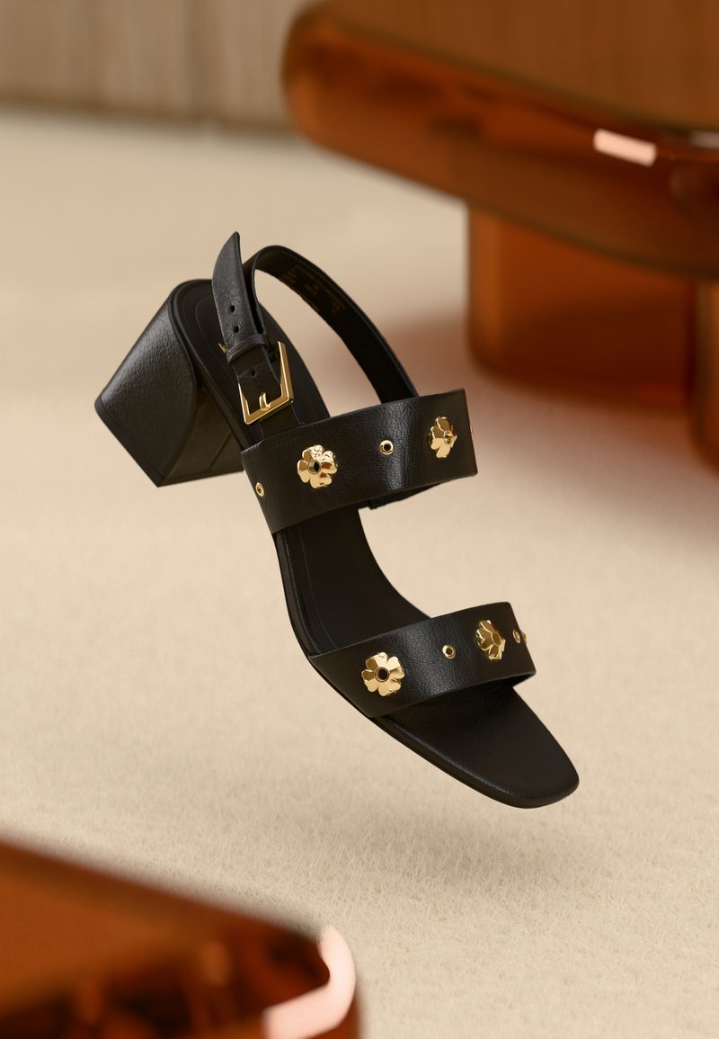 Black leather heeled sandals with two straps featuring gold flower-shaped embellishments and a buckle. Square heel design. Smooth texture.