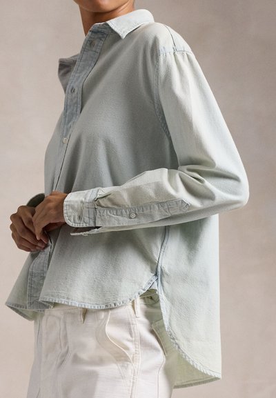 Személy, aki világoskék hosszú ujjú gombos inget és fehér nadrágot visel, kezét a dereka mellett összefonva, egyszínű háttér előtt.