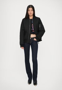 Black quilted bomber jacket with zippered pocket, worn over a cropped multicolored knit top, paired with dark blue flared jeans and black heels.