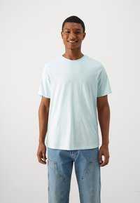 Camiseta verde menta clara hecha de tela suave. Cuenta con un cuello redondo y mangas cortas. Combinada con jeans azules, de pie contra un fondo neutro.