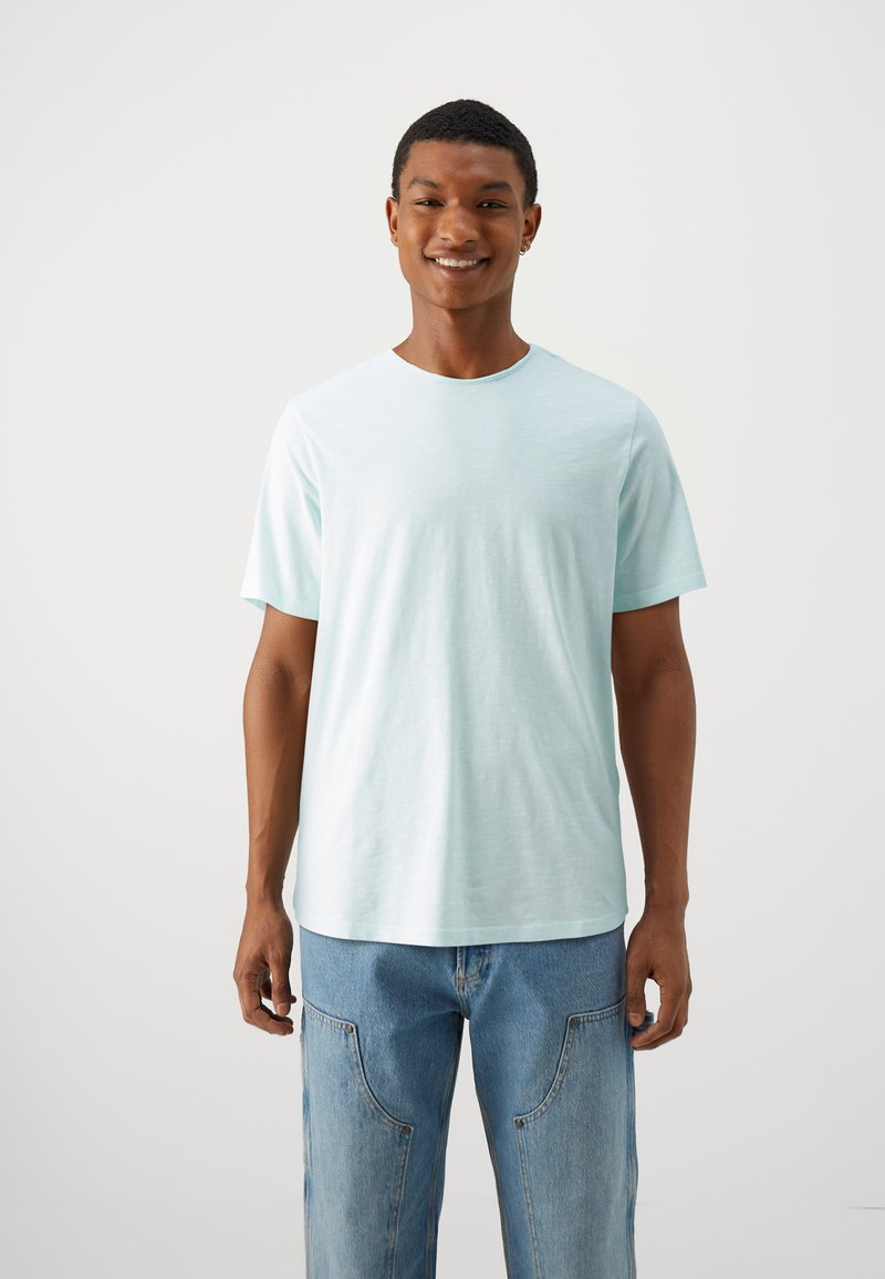 Camiseta verde menta clara hecha de tela suave. Cuenta con un cuello redondo y mangas cortas. Combinada con jeans azules, de pie contra un fondo neutro.