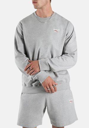 Mann trägt hellgraues Reebok-Sweatshirt und passende Shorts, die Hände verschränkt, mit silbernen Ringen und einer Kettenhalskette, vor schlichtem Hintergrund.