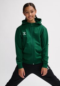 Grön zip-up hoodie med ficka fram, tillverkad av ett mjukt material. Har en huva och en vit logotyp på vänster bröst. Matchas med svarta byxor.