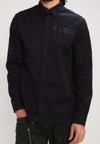 Man som bär en svart långärmad skjorta med knappar och krage samt mörka byxor med dragkedjefickor.