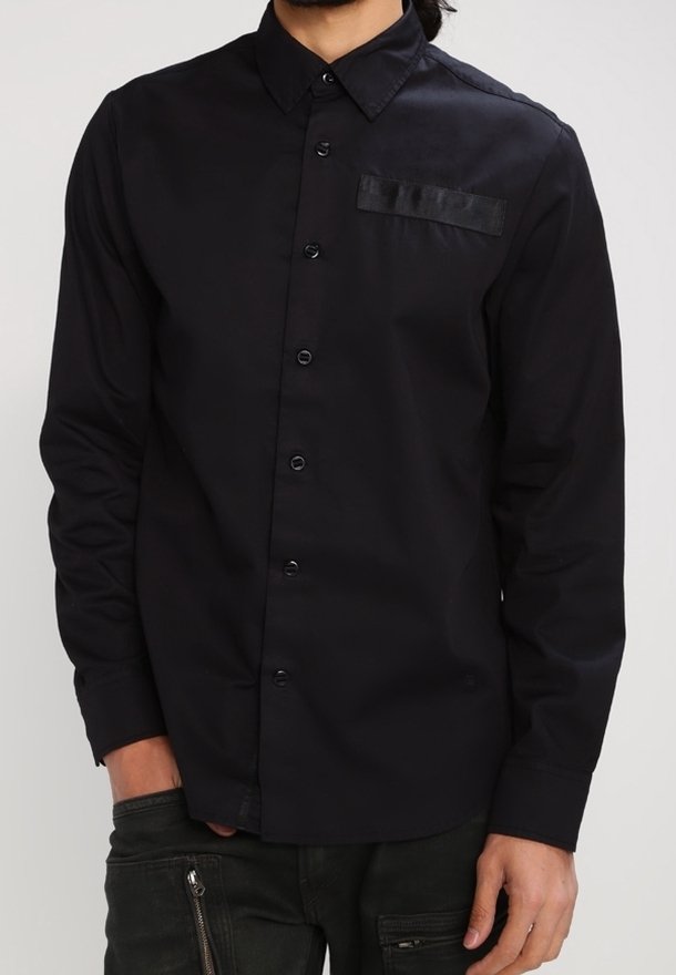 Man som bär en svart långärmad skjorta med knappar och krage samt mörka byxor med dragkedjefickor.