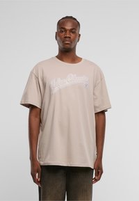 Beige bomull T-skjorte med avslappet passform, som har lyst lilla "Urban Classics" tekst. Korte ermer og rund halslinning uten synlige mønstre.