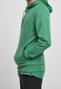 Grüner Kapuzenpullover aus weichem Stoff, mit einer Fronttasche und verstellbarem Kordelzug an der Kapuze. Details sind gerippte Bündchen und Saum.