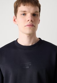 Fekete pulóver kerek nyakkivágással, matt "PORSCH BOSS" felirattal a mellkasán. Simaságot idéző, pamutszerű textúra.