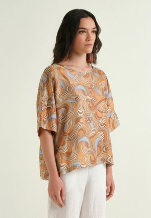 Donna con capelli scuri fino alle spalle indossa una maglia larga con motivi arancioni, blu e crema e pantaloni bianchi, rivolta leggermente a destra con espressione neutra.