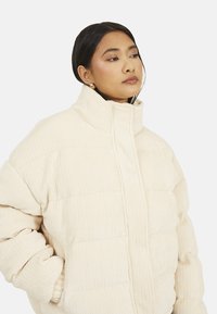 Kurtka puchowa w kolorze ecru z pionową, prążkowaną fakturą, wysokim kołnierzem i luźnym krojem. Wyposażona w zapięcie na suwak oraz boczne kieszenie.