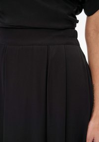 Detail van een zwarte jurk met zachte stof, plooien bij de taille en een elastische tailleband. Ontwerp benadrukt een gladde, vloeiende silhouet.