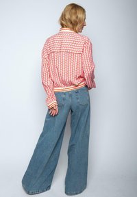 Bunte Bomberjacke mit einem geometrischen Muster in Rot, Orange und Weiß, kombiniert mit weit geschnittenen blauen Jeans, von hinten betrachtet.