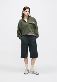 Νέος με καστανά μαλλιά που φοράει ένα ελαιόχρωμο κοντό φούτερ με φερμουάρ, μαύρες σορτς μέχρι το γόνατο, κρεμ κάλτσες και λευκά αθλητικά παπούτσια.